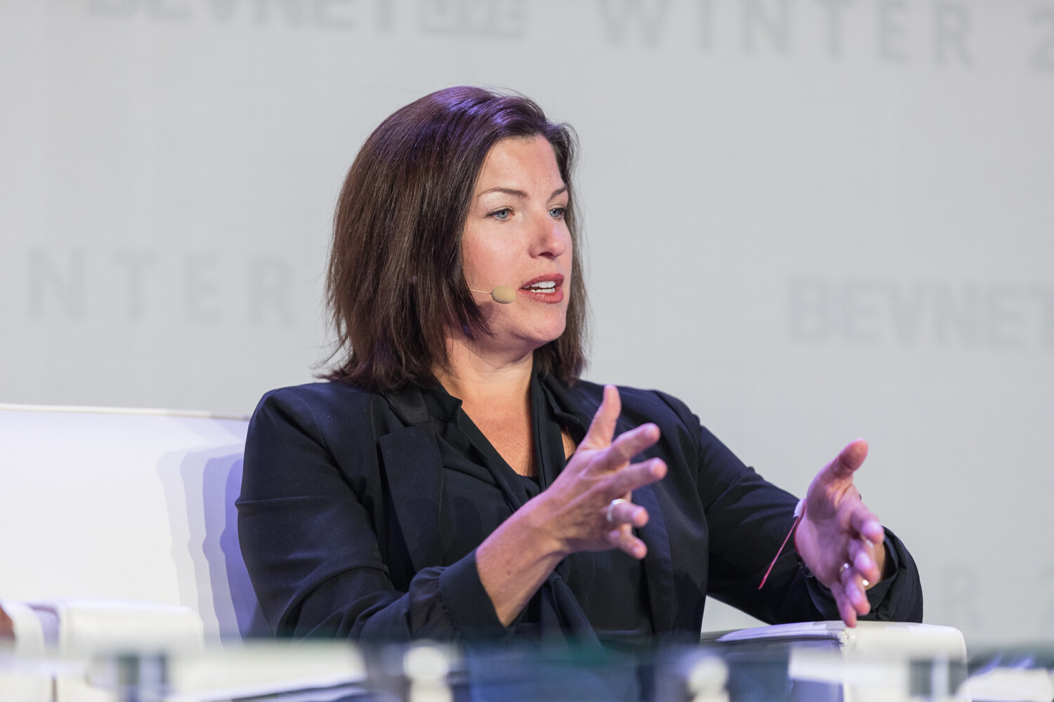 A woman with shoulder-length brown hair in a black jacket and headset microphone gestures with both hands while speaking.