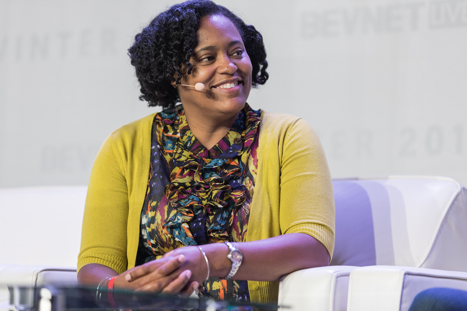 A smiling woman with curly dark hair, a yellow cardigan and a colorful ruffled blouse sits on a white sofa wearing a headset microphone.