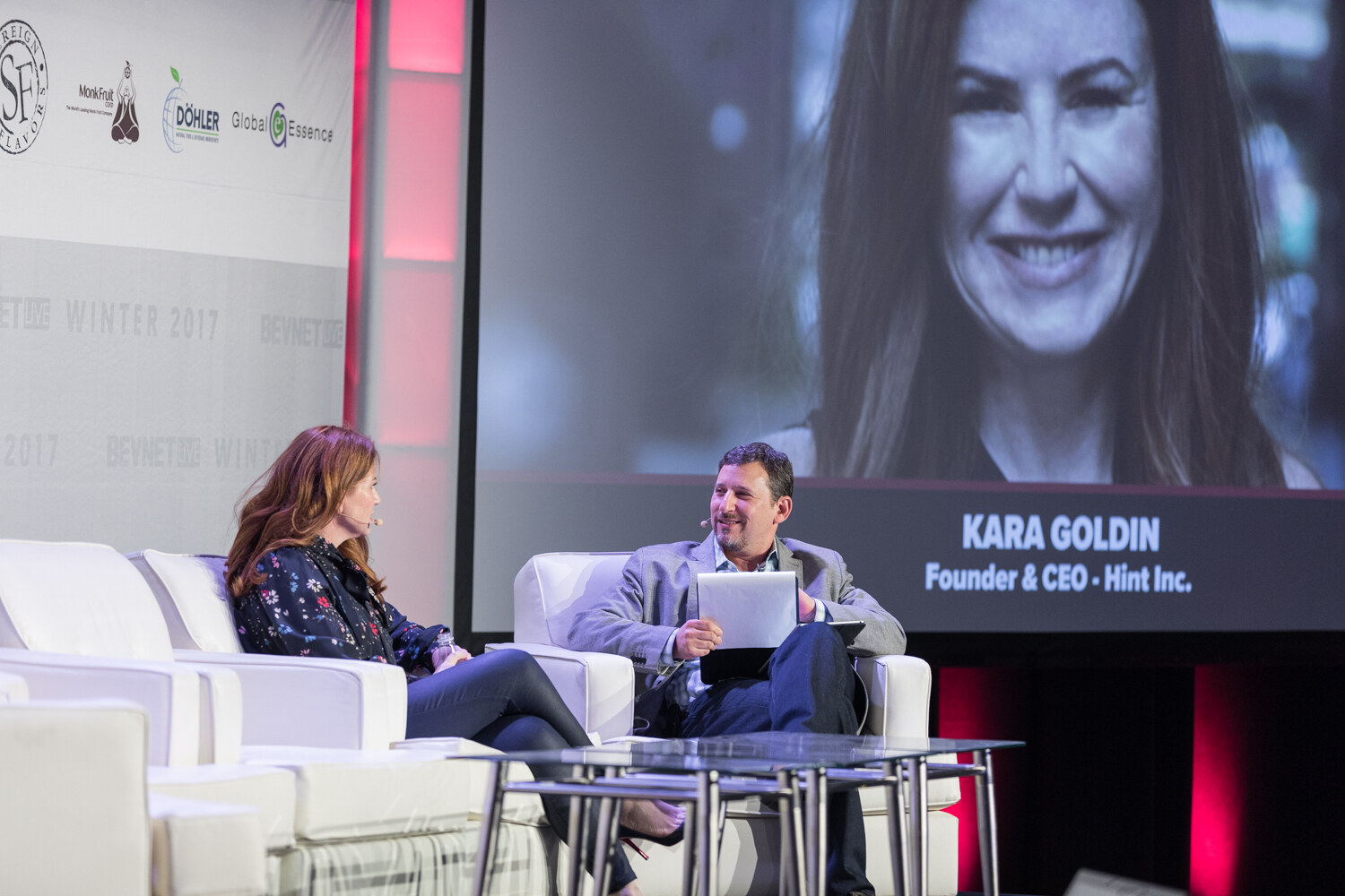 A woman with auburn hair and a man in a gray blazer holding a tablet sit on white sofas, with a large screen behind them showing a smiling woman and the caption Founder and CEO, Hint Inc.