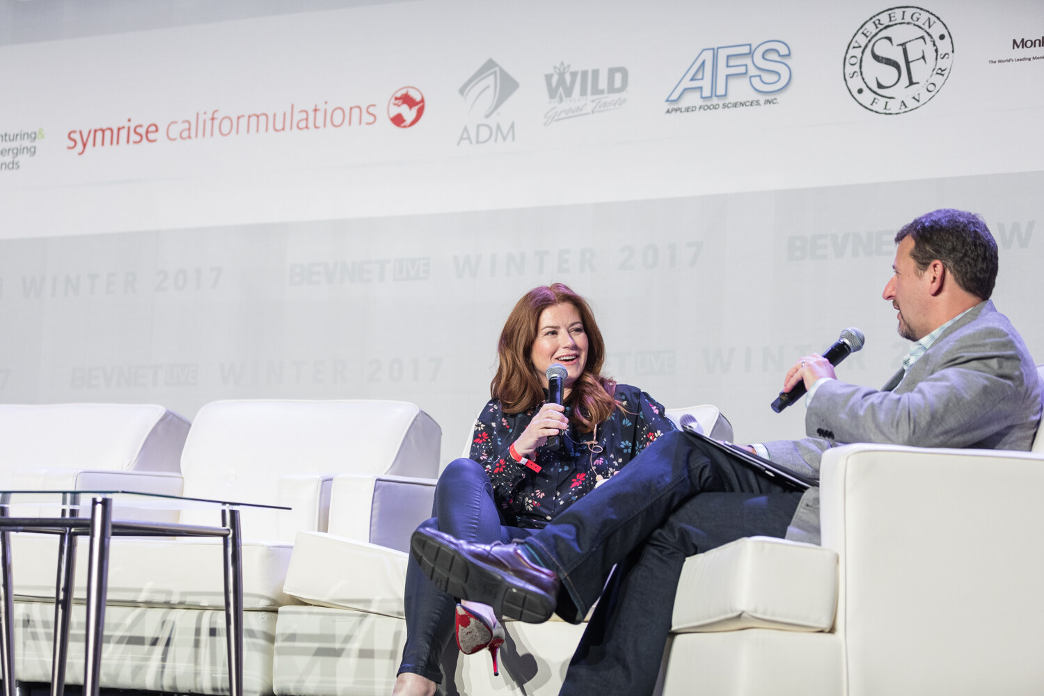 A woman in a dark floral blouse and red heels holds a microphone and laughs while a man in a gray blazer with a handheld microphone and tablet sits across from her.