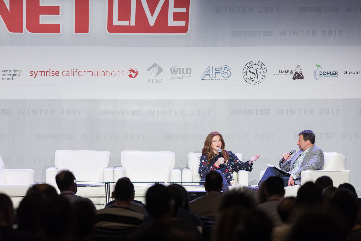 Wide view over the heads of the audience of a woman and a man seated on white sofas on a stage with BevNET Live sponsor banners.