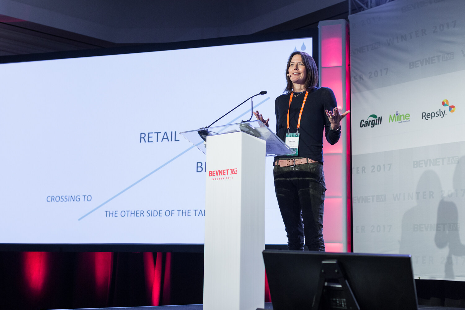 A woman in a black top with an orange lanyard stands at a white lectern with both hands raised, beside a slide that reads Retail, Crossing to the other side of the table.