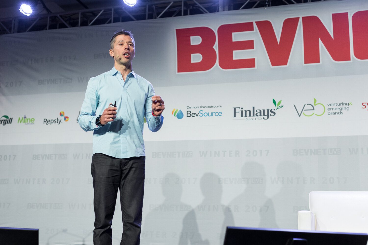 A man in a light blue button-up shirt and dark pants holds a slide clicker and speaks on a stage in front of a BevNET Live banner with sponsor logos.