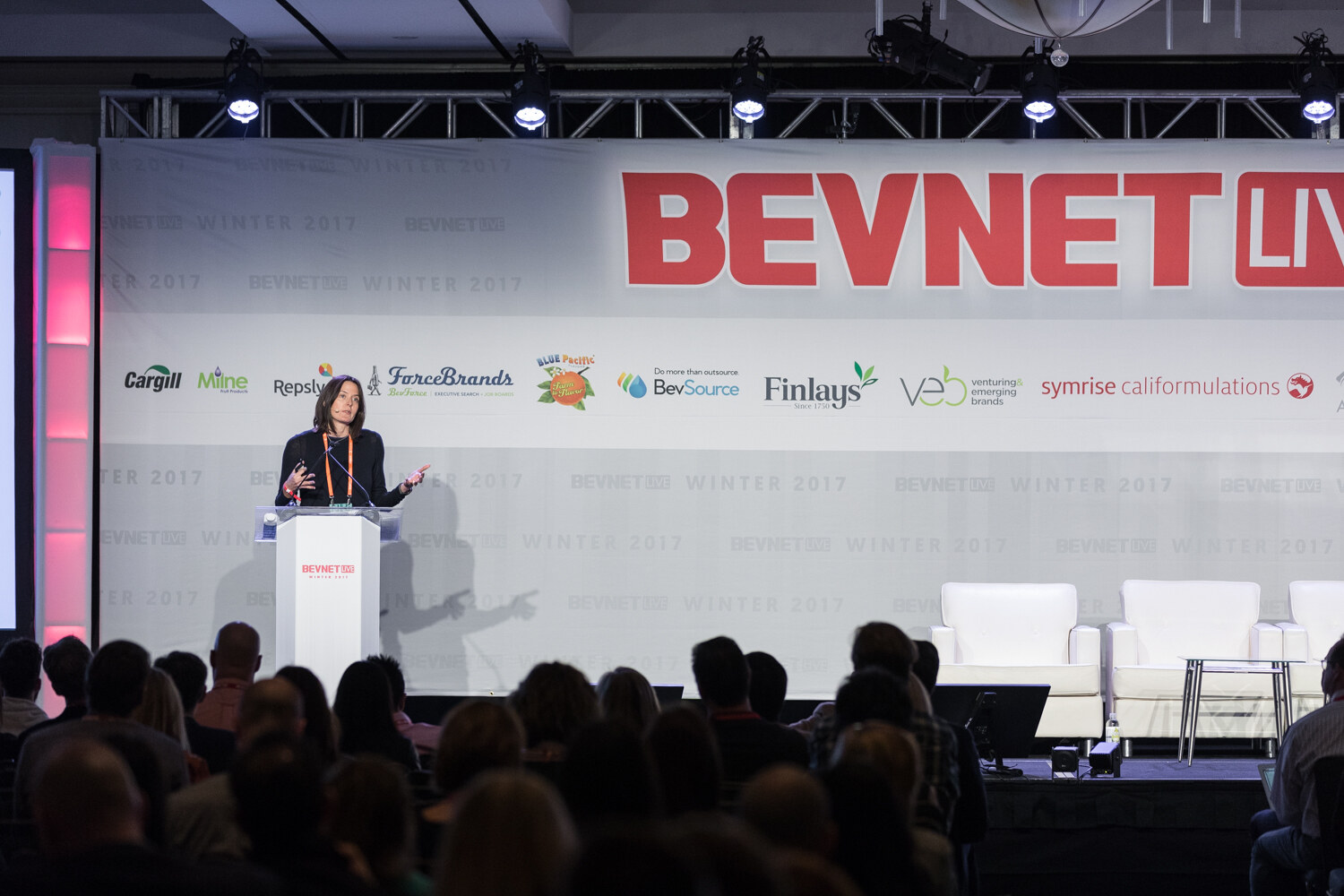 A woman in black stands at a white lectern in front of a large BevNET Live banner with sponsor logos, seen over the heads of the audience.