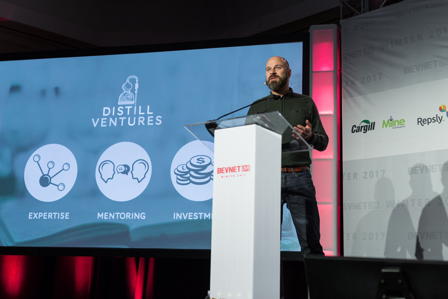 A bald bearded man in a dark green shirt speaks at a white lectern beside a blue slide with the Distill Ventures logo and icons labeled Expertise, Mentoring and Investment.
