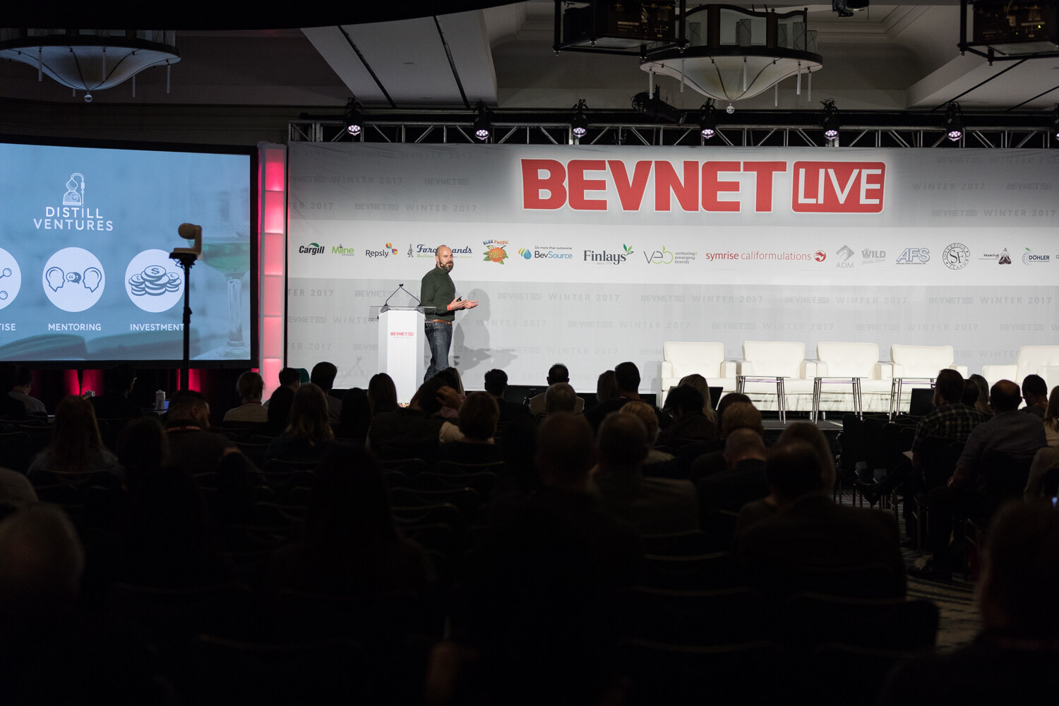 Wide view over the audience of a man at a lectern under a large BevNET Live banner, with a screen at left showing the Distill Ventures logo and icons.