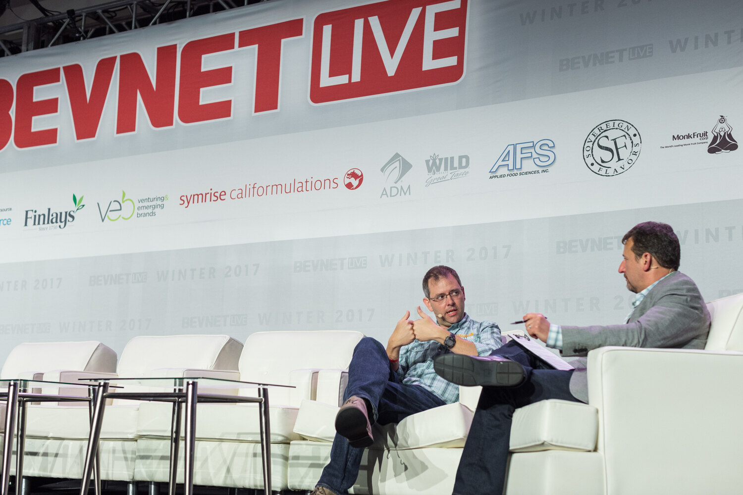 Two men on white sofas on a stage under the BevNET Live banner, one in a light plaid shirt and glasses gesturing, the other in a gray blazer writing on a notepad.