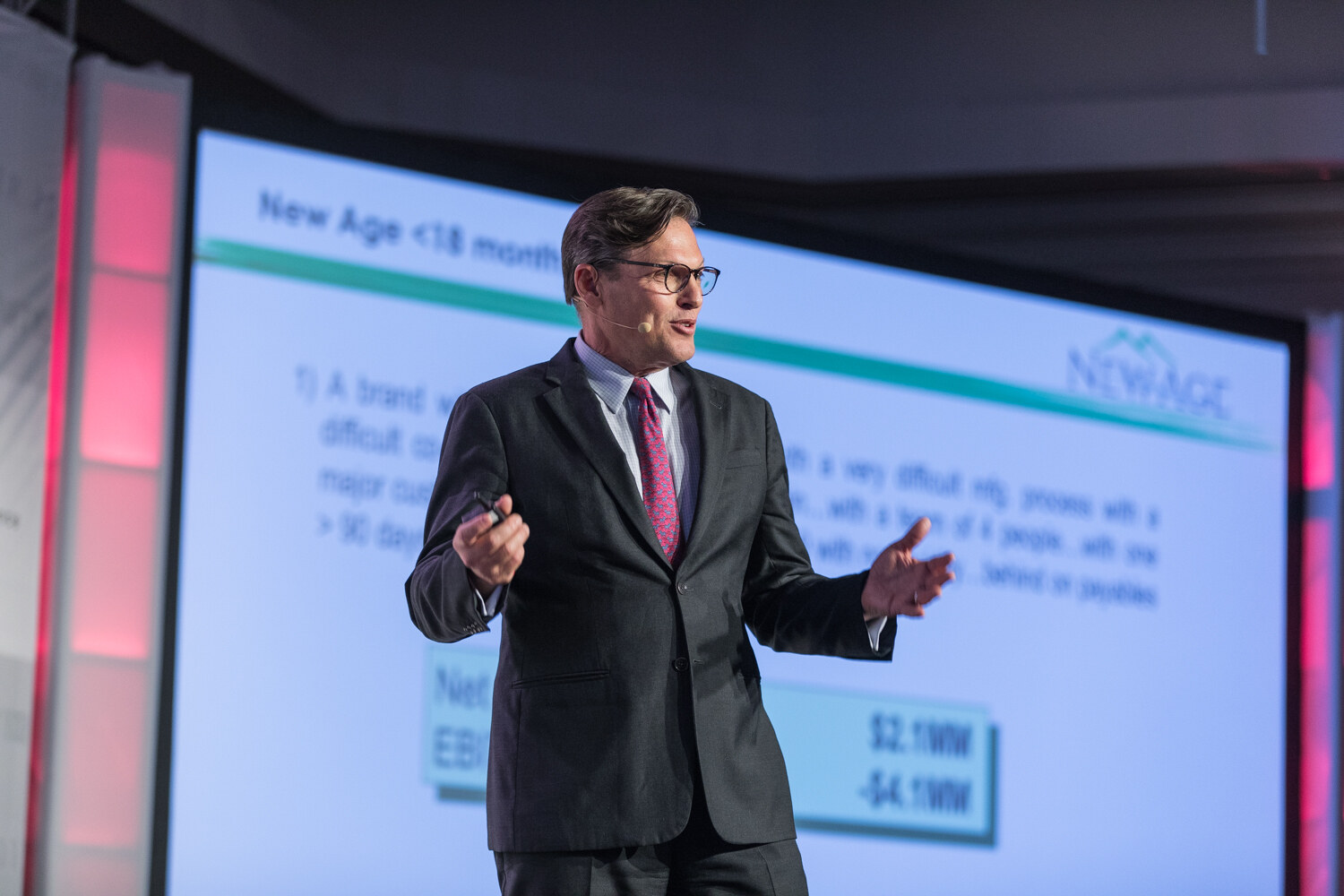 A man in a dark suit, glasses and magenta tie holds a clicker and gestures in front of a slide headed New Age less than 18 months ago with the New Age logo.