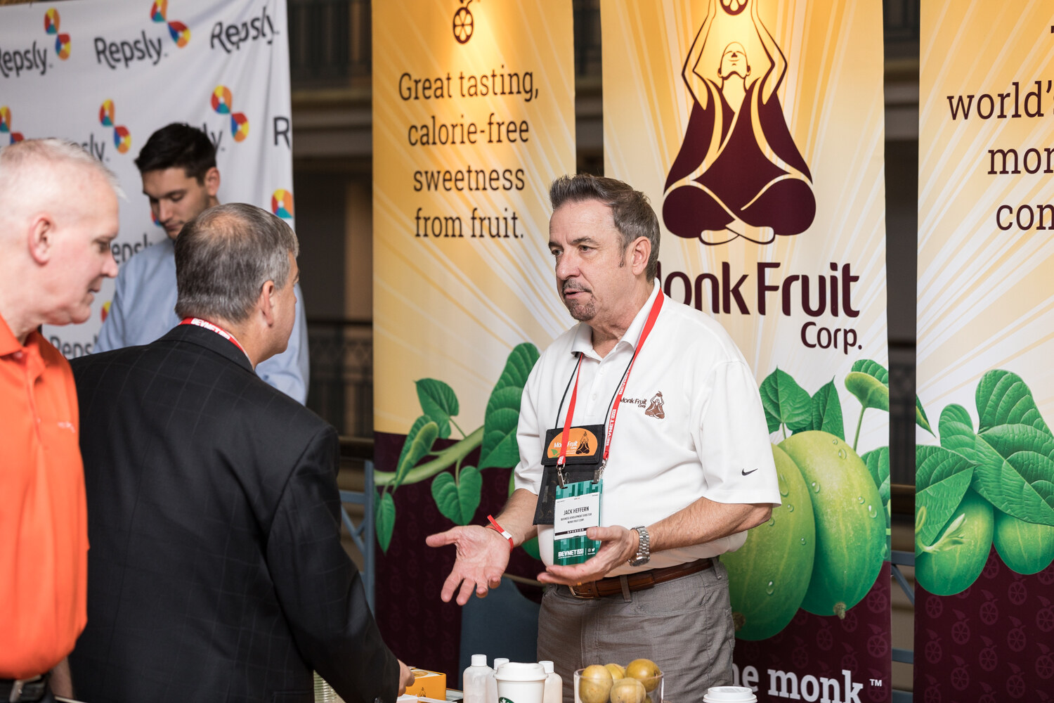 A man in a white polo with a conference lanyard gestures while talking with a visitor at a booth with Monk Fruit Corp. banners reading Great tasting, calorie-free sweetness from fruit, with Repsly banners behind.