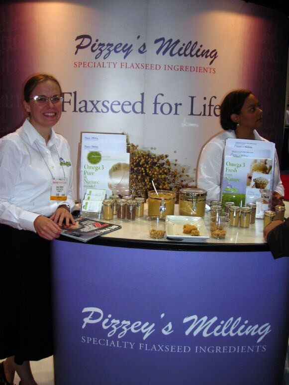 Two women in white shirts stand behind a curved purple Pizzey's Milling booth counter with jars of flaxseed ingredients, brochures and a Flaxseed for Life backdrop.