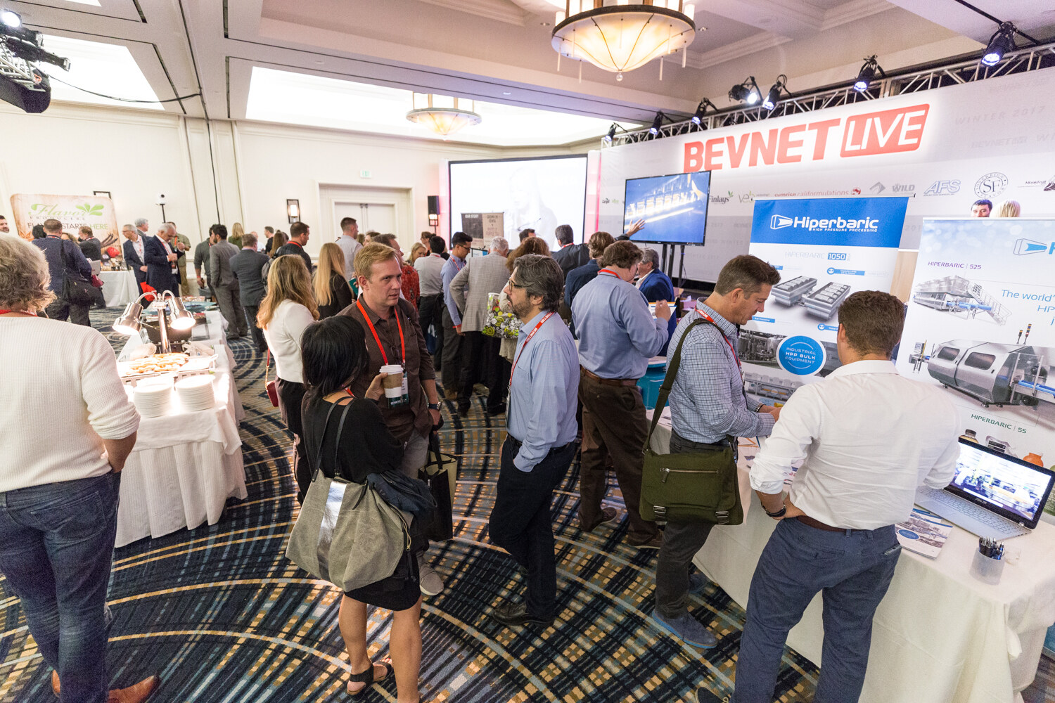 Wide view of a ballroom expo floor with attendees at a Hiperbaric high-pressure processing display, a buffet table and a BevNET Live banner overhead.