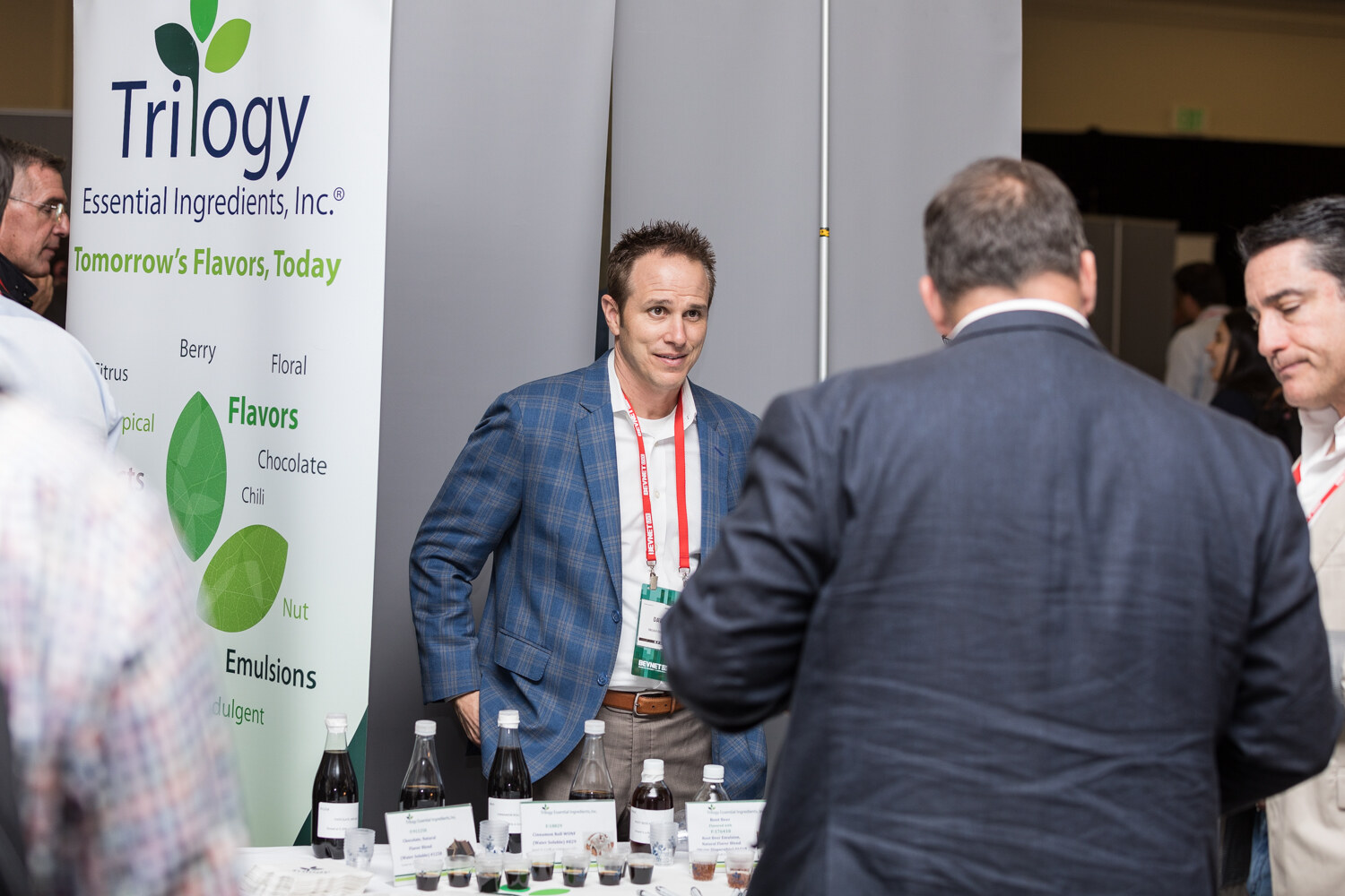 A man in a blue plaid blazer with a red lanyard stands at a booth with a Trilogy Essential Ingredients banner and rows of small flavor sample bottles and cups, as visitors talk in the foreground.