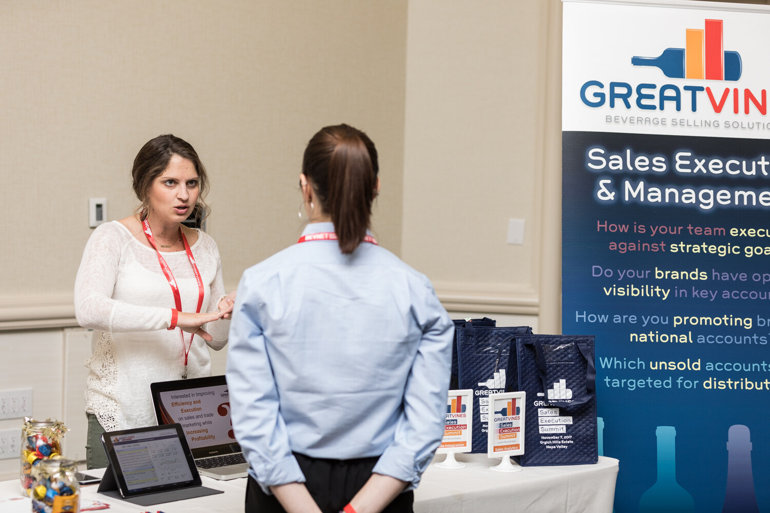 A woman in a white lace top with a red lanyard talks with a visitor in a blue shirt at a table with laptops and branded bags in front of a GreatVines banner about sales execution and management.