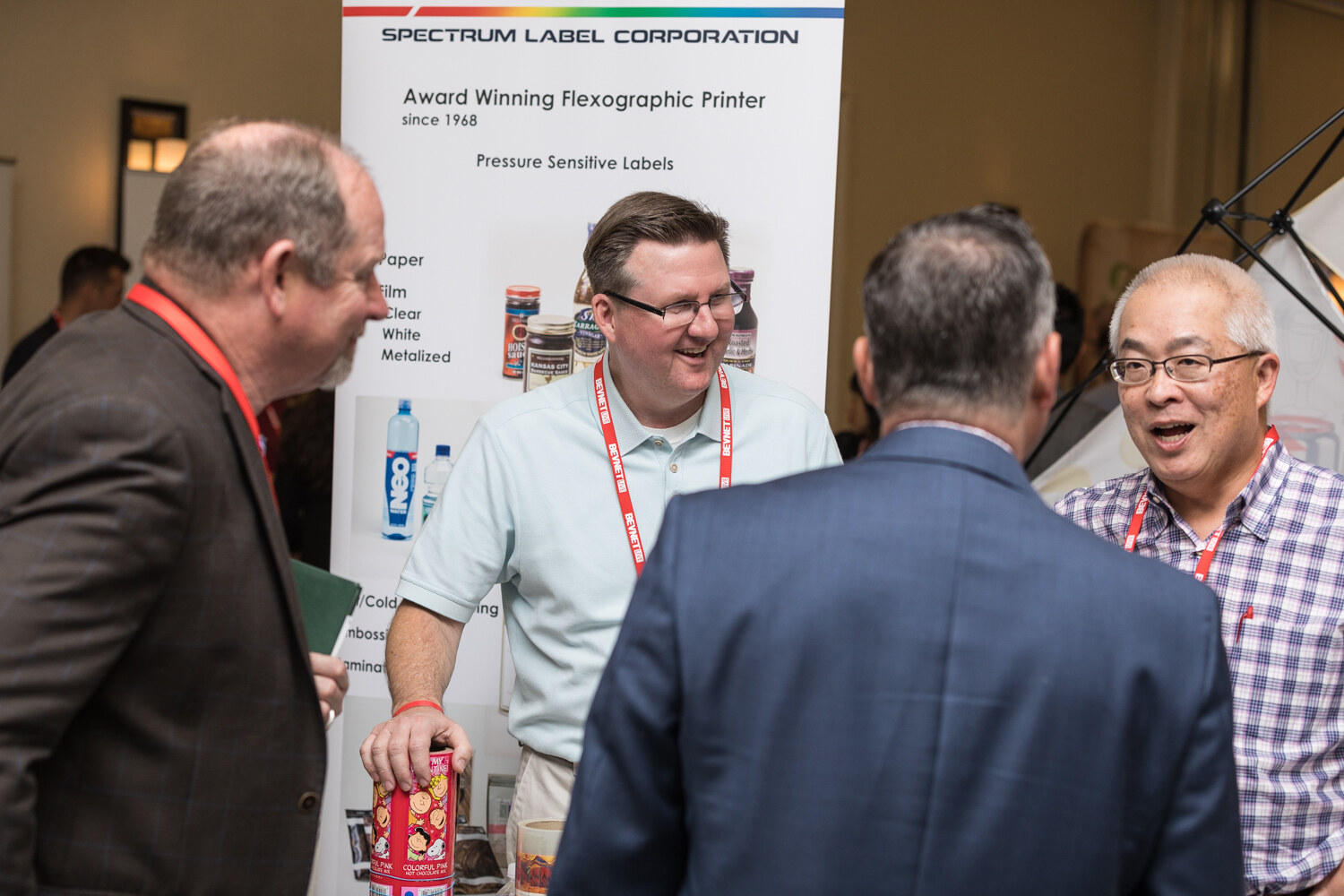 Four men chat and laugh at a booth in front of a Spectrum Label Corporation banner advertising pressure sensitive labels, one holding a decorated can.