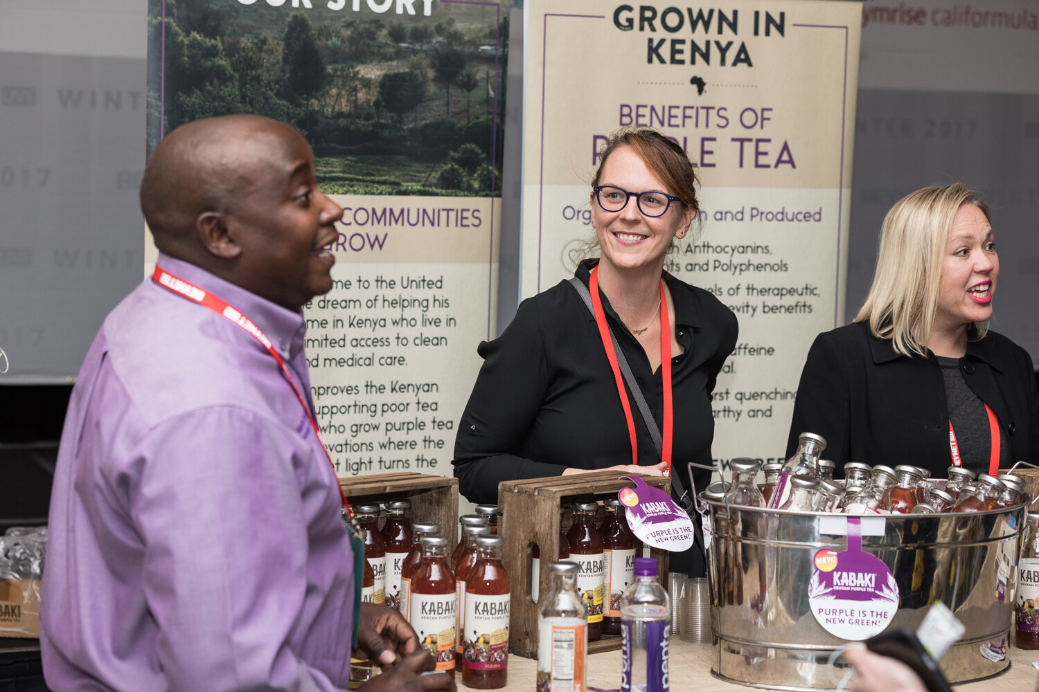 A man in a purple shirt and two smiling women stand behind a bucket and crates of Kabaki tea bottles in front of banners reading Grown in Kenya and Benefits of Purple Tea.
