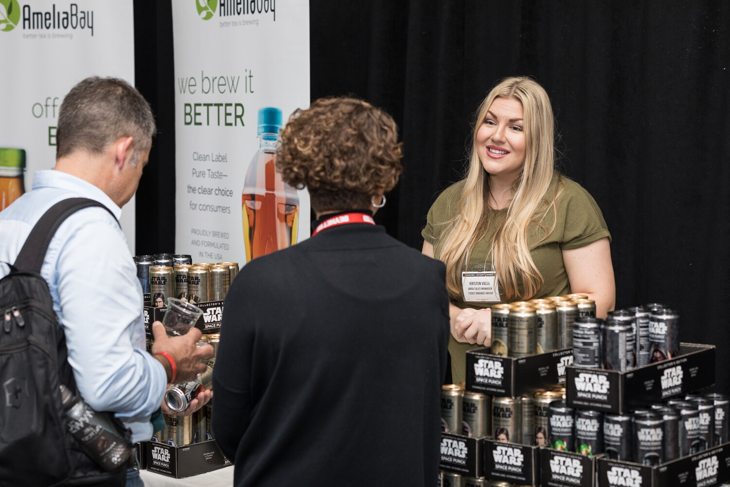 A woman with long blonde hair in an olive T-shirt smiles at two visitors behind stacked boxes of Star Wars Space Punch cans, in front of AmeliaBay banners reading We brew it better.