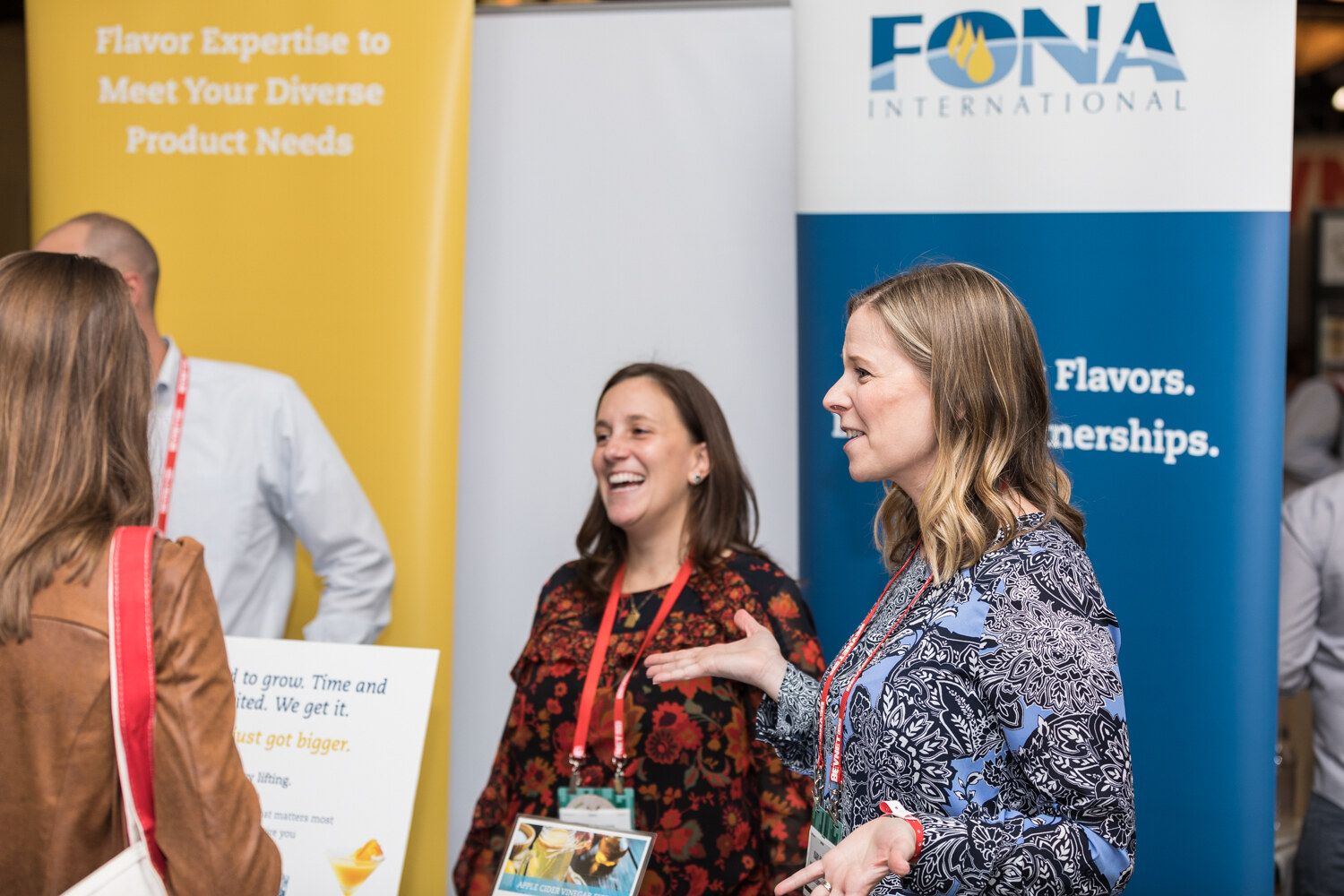 Two women with red lanyards laugh and talk beside a FONA International banner reading Flavors and Partnerships, with a visitor in a brown jacket in the foreground.