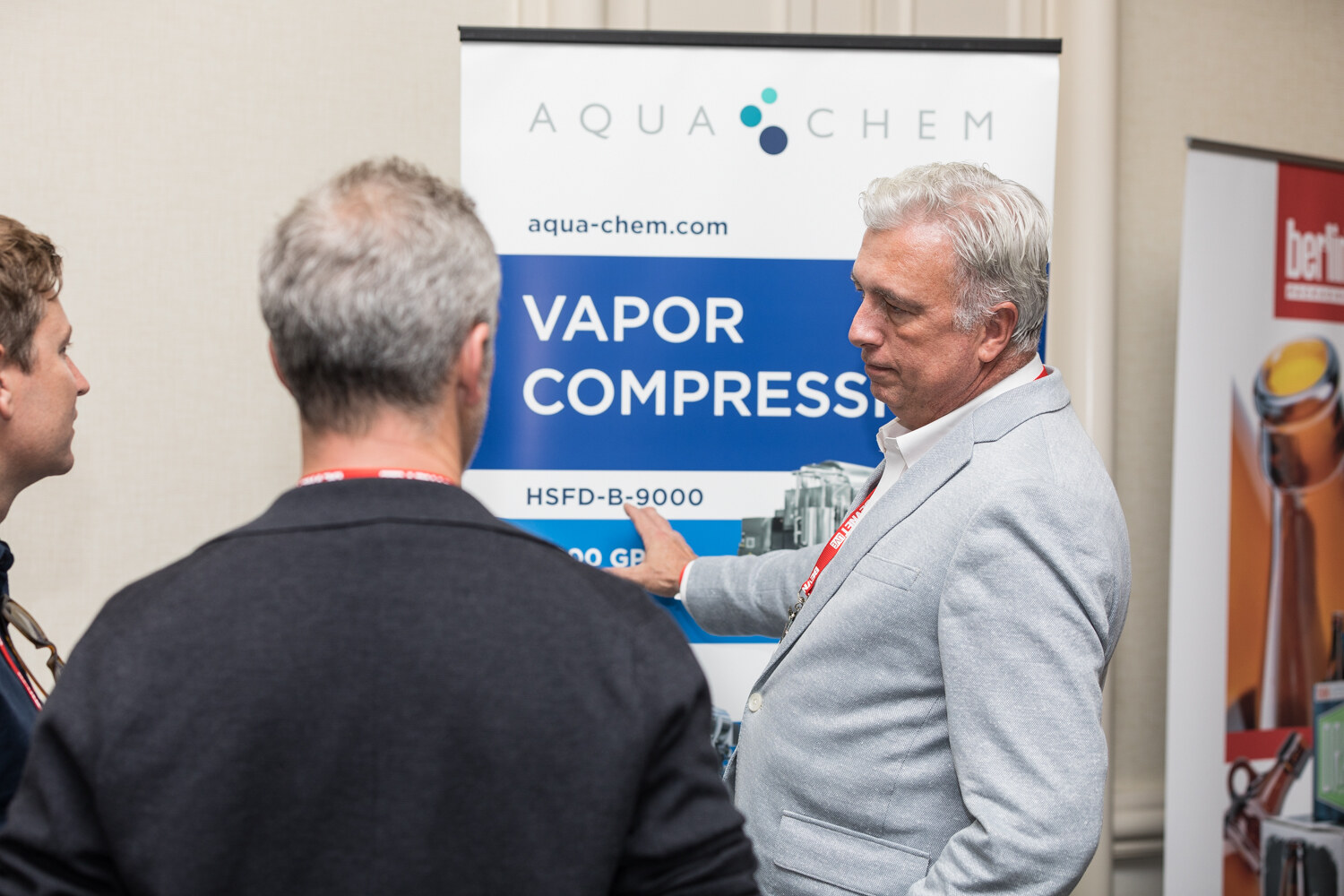 A gray-haired man in a light gray blazer points to an Aqua-Chem banner reading Vapor Compression while two visitors listen, with a Berlin Packaging banner at right.