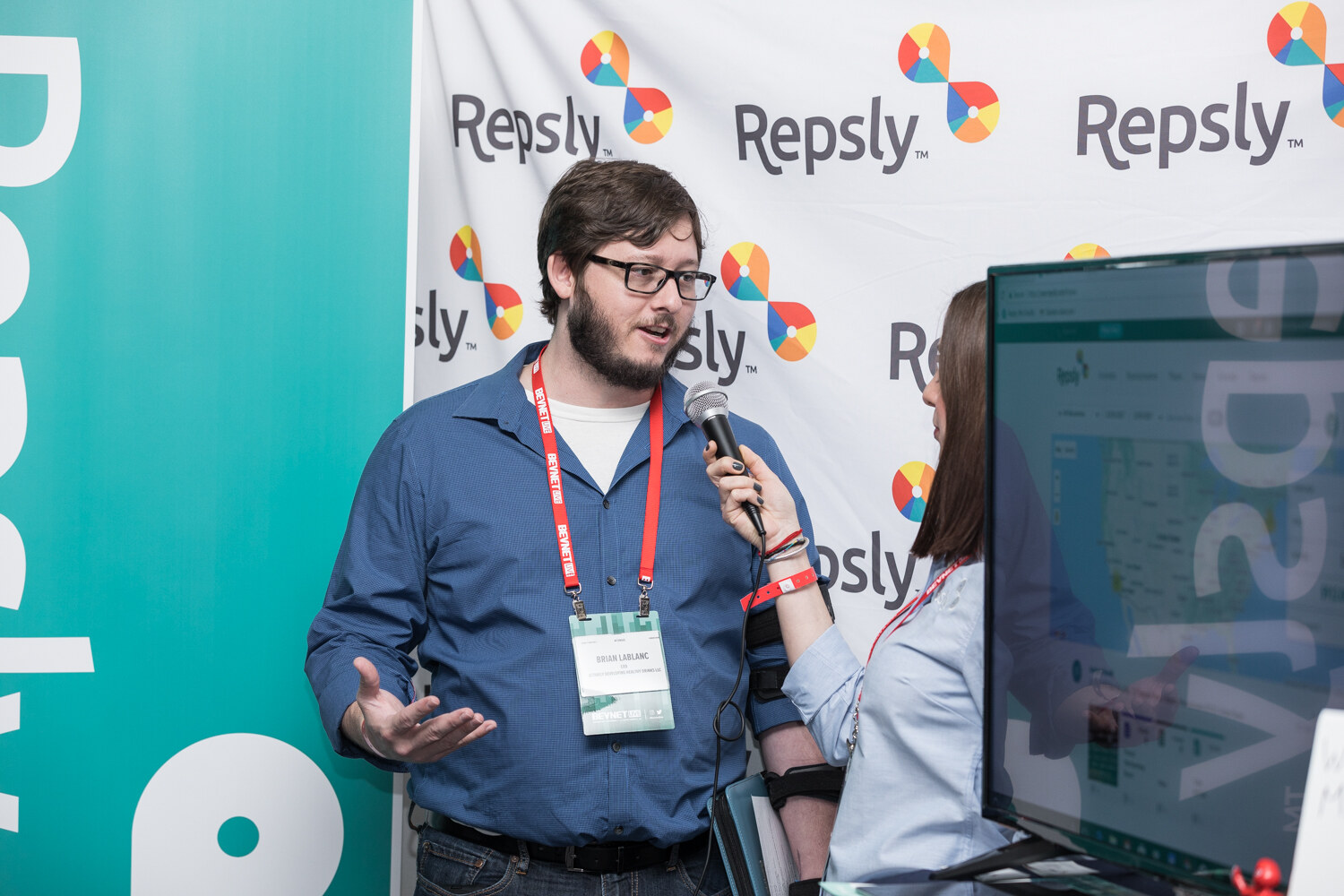 A bearded man in glasses and a blue shirt gestures while a woman holds a microphone toward him in front of a Repsly step-and-repeat banner and a monitor.