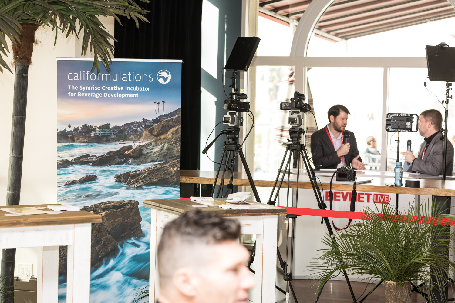 Two men sit at a counter with a BevNET Live front panel being filmed by cameras on tripods, next to a califormulations banner about a creative incubator for beverage development and a potted palm.