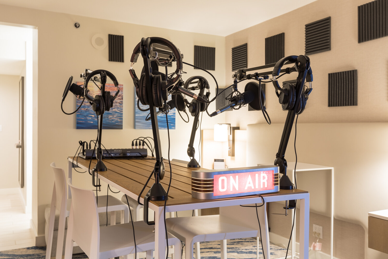 A wooden table with four boom-arm microphones, headphones, an audio interface and a glowing On Air sign in a hotel room lined with acoustic foam panels.