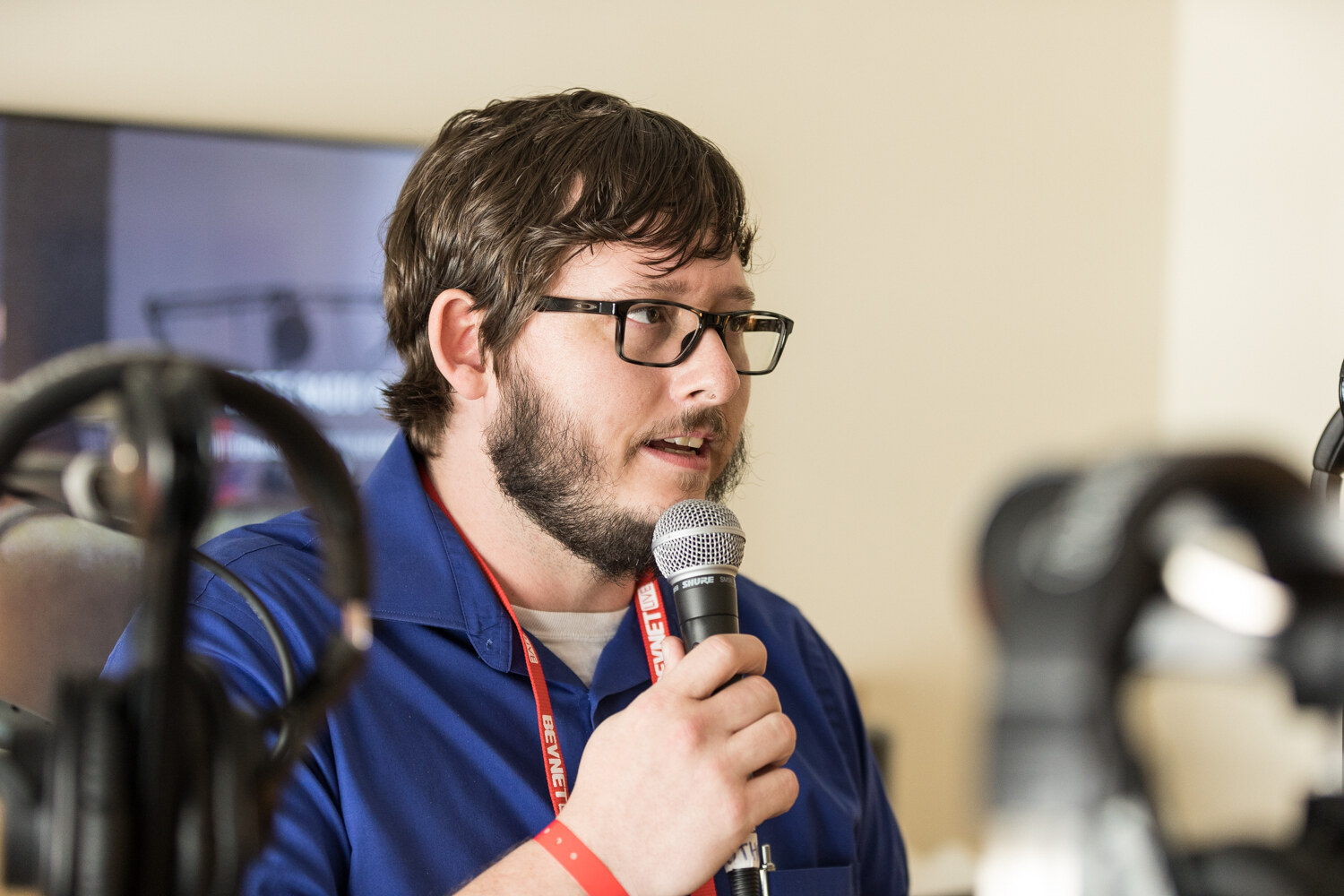 A bearded man in glasses and a blue button-up shirt with a red lanyard speaks into a handheld microphone, with headphones on stands blurred in the foreground.