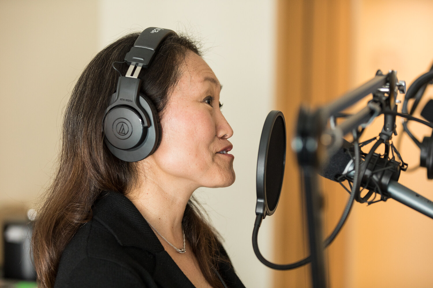 Profile of a woman with long dark hair wearing over-ear headphones, speaking toward a studio microphone with a round pop filter.