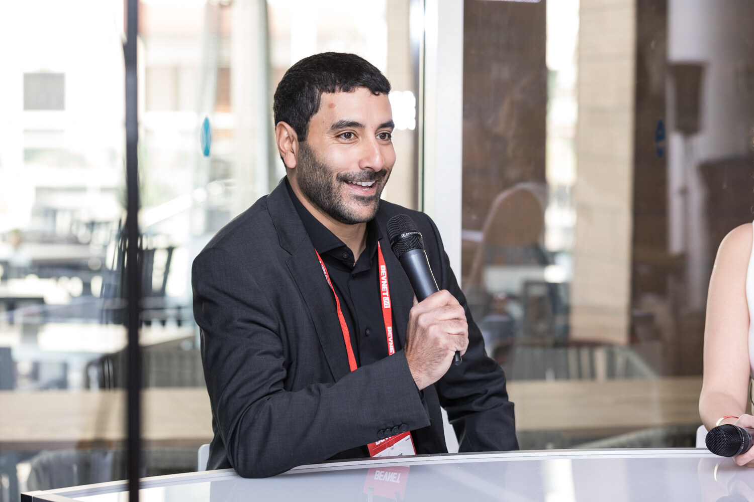 A smiling man with a short beard in a dark blazer and red BevNET lanyard holds a handheld microphone at a white table in front of a glass wall.