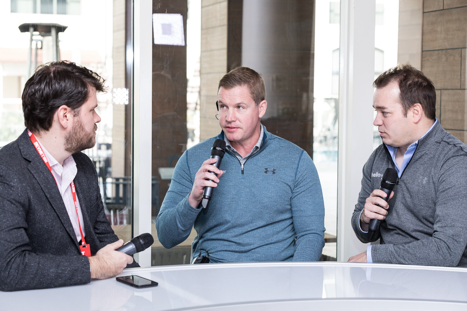 Three men hold handheld microphones at a round white table in front of glass windows, one in a dark blazer and red lanyard, one in a blue Under Armour pullover and one in a gray pullover.