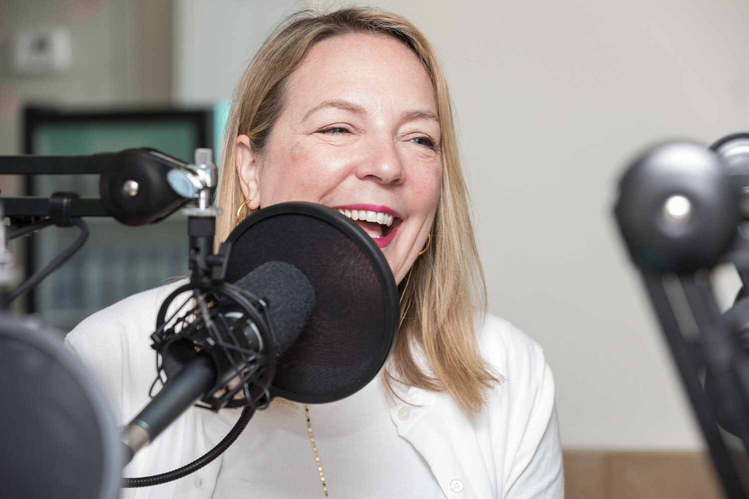 A smiling blonde woman in a white cardigan laughs behind a studio microphone with a black pop filter in a recording room.