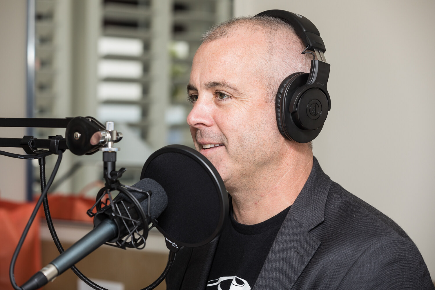 Close-up of a man with closely cropped hair wearing headphones and a dark blazer, speaking beside a studio microphone with a pop filter.