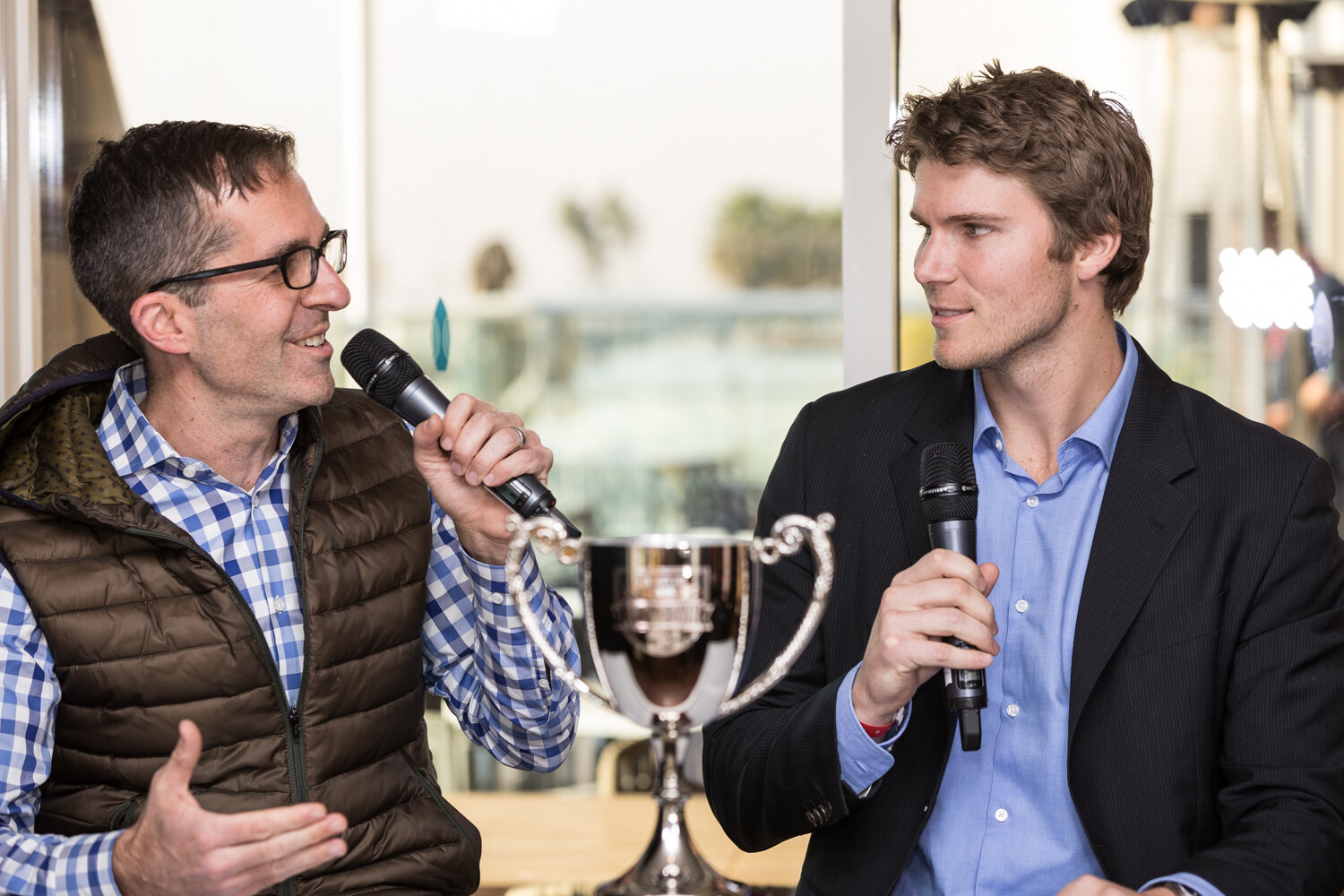 Two men hold handheld microphones and talk, one in a brown puffer vest and blue checked shirt, the other in a navy blazer, with a silver trophy cup between them in front of a window.