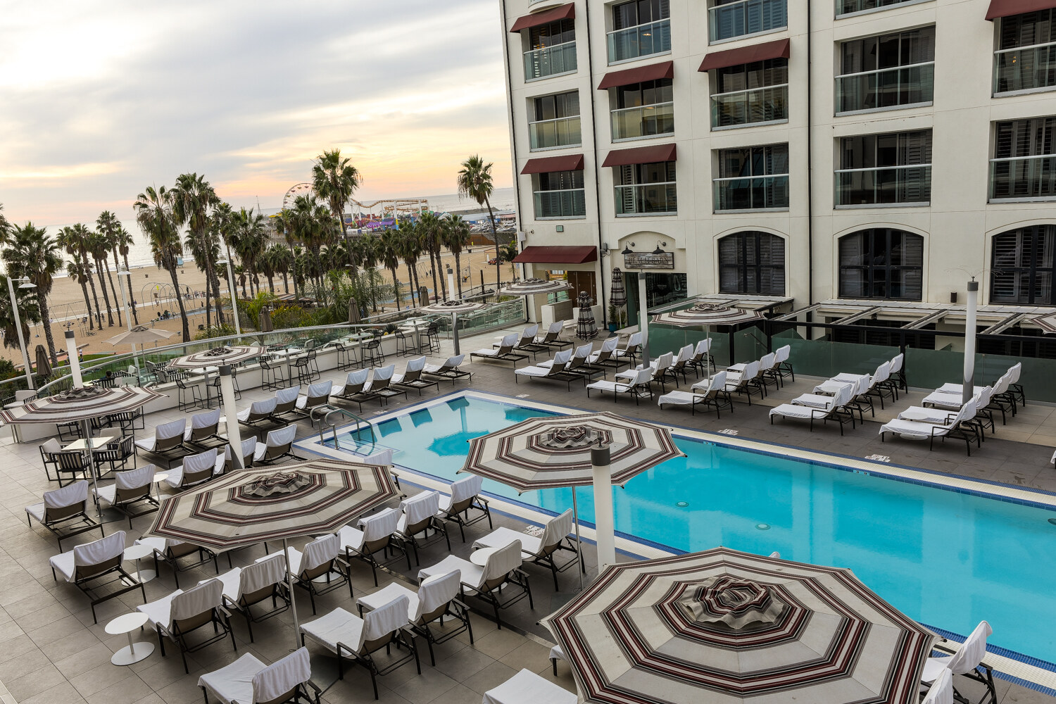 Elevated view of a hotel pool deck with striped umbrellas, white lounge chairs and a blue pool, with palm trees, the beach and a Ferris wheel on the pier under a pastel sky.