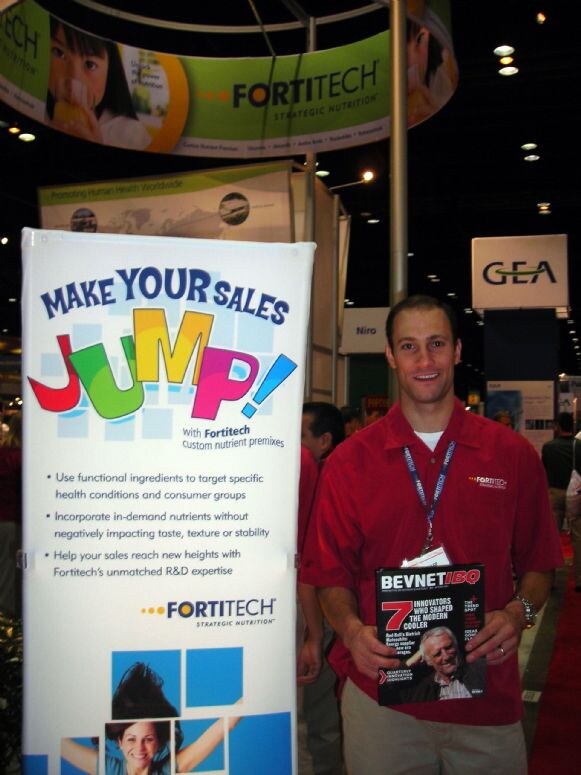 A smiling man in a red polo and lanyard holds a BevNET magazine beside a Fortitech banner reading "Make your sales jump!" A Fortitech sign hangs above and a GEA sign is behind him.