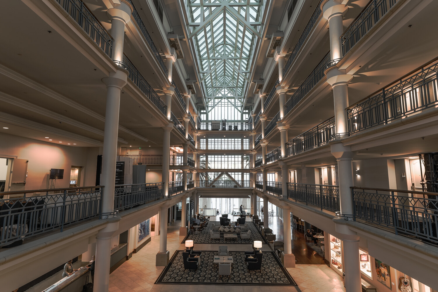 View down the skylit atrium of a hotel with white columns, wrought-iron balconies and a lobby seating area with patterned rugs at the bottom.