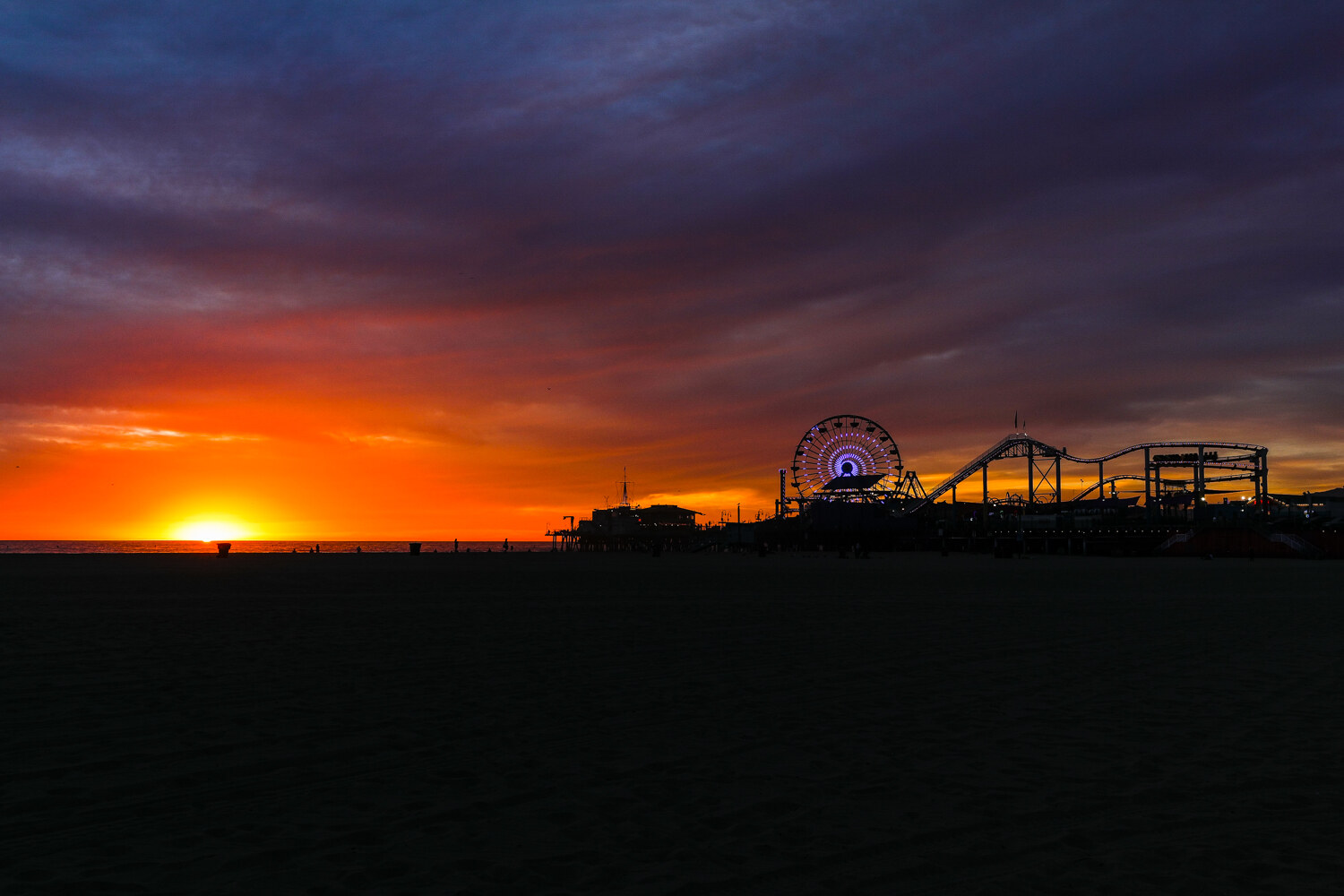 A vivid orange and purple sunset over the ocean with the silhouettes of the Santa Monica Pier Ferris wheel and roller coaster on the right.