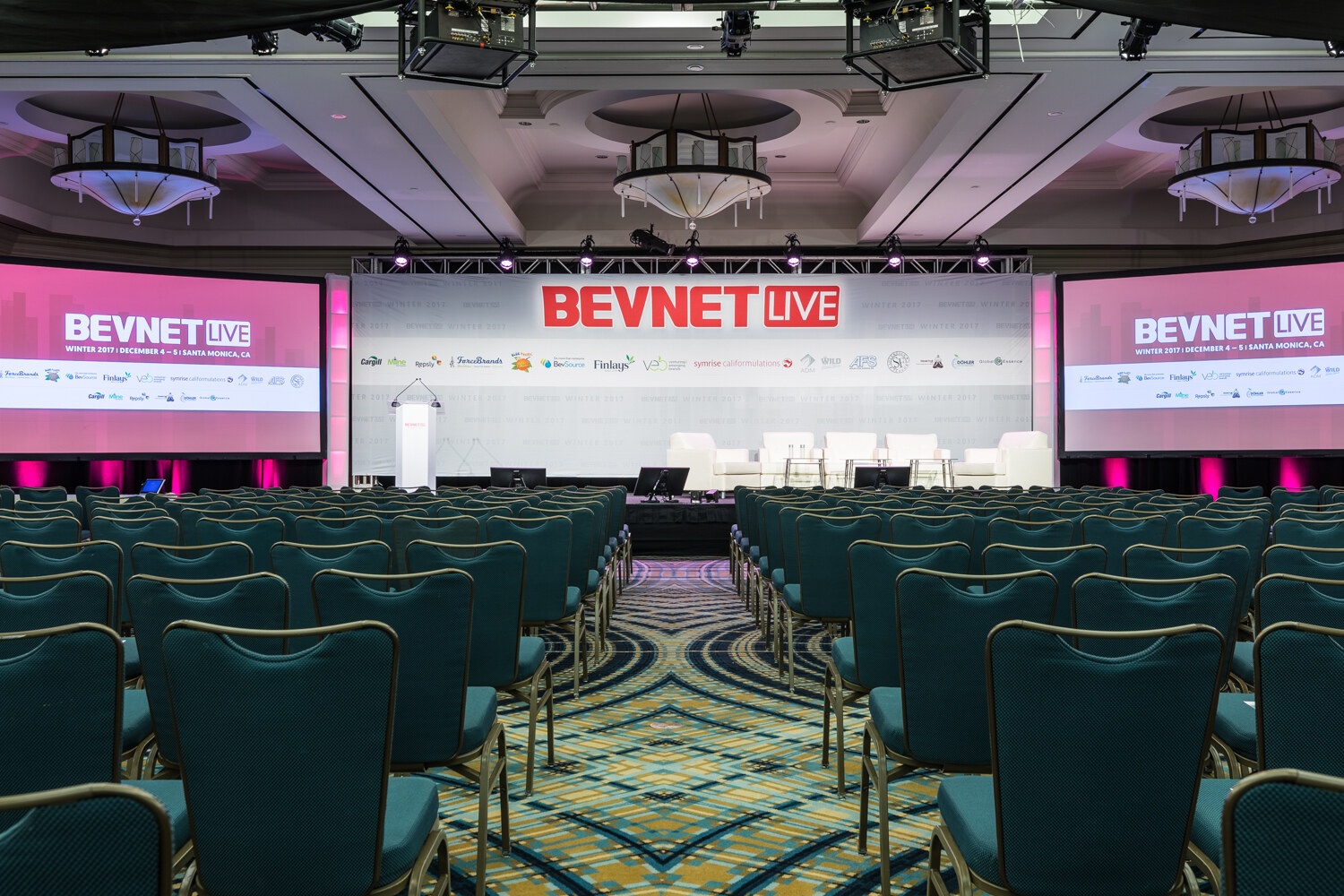 Wide view down an aisle of teal chairs toward a stage with white sofas, a white lectern and screens on either side reading BevNET Live Winter 2017, December 4-5, Santa Monica, CA.