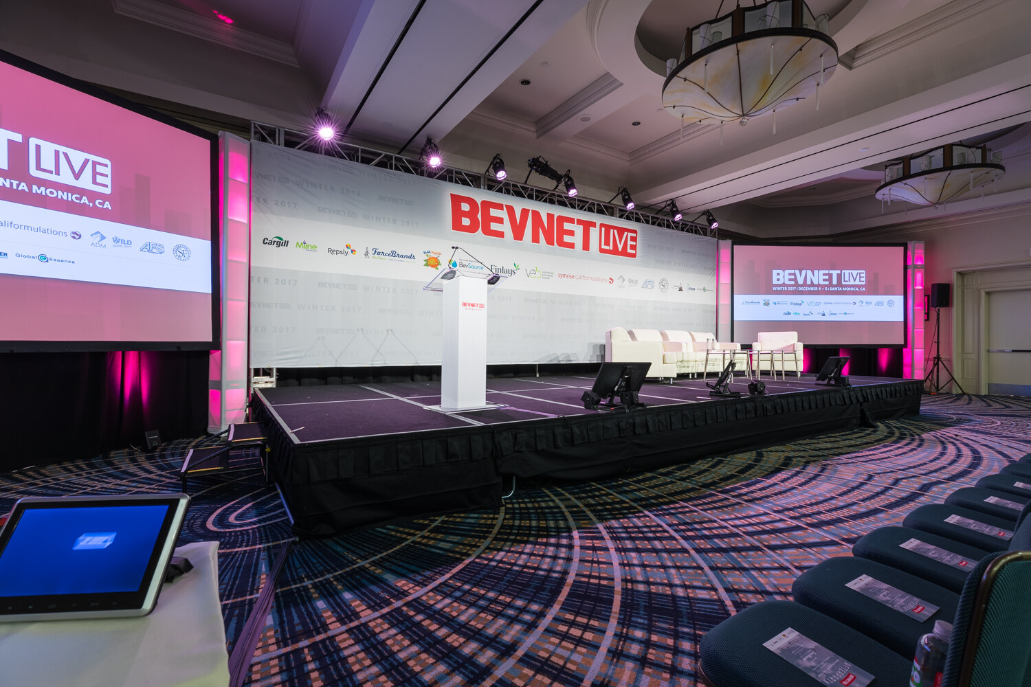 Low-angle view of a black-skirted stage with a white lectern, white sofas and a BevNET Live backdrop with sponsor logos, with a floor monitor and chairs in the foreground.