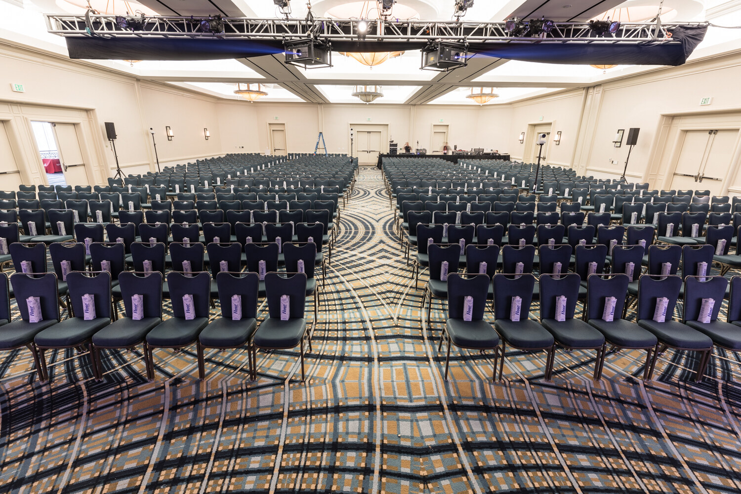 Wide view of a large ballroom filled with rows of navy chairs, each with a small purple carton on the seat, facing a stage far ahead.