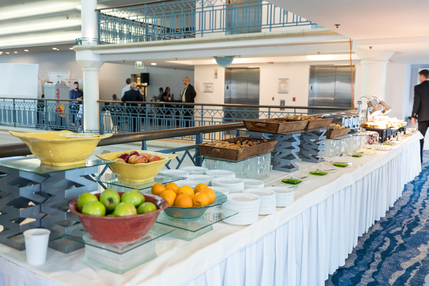 A long white-skirted buffet table with bowls of green apples and oranges, plates, pastries and chafing dishes on the hotel mezzanine, with elevators and balconies behind.