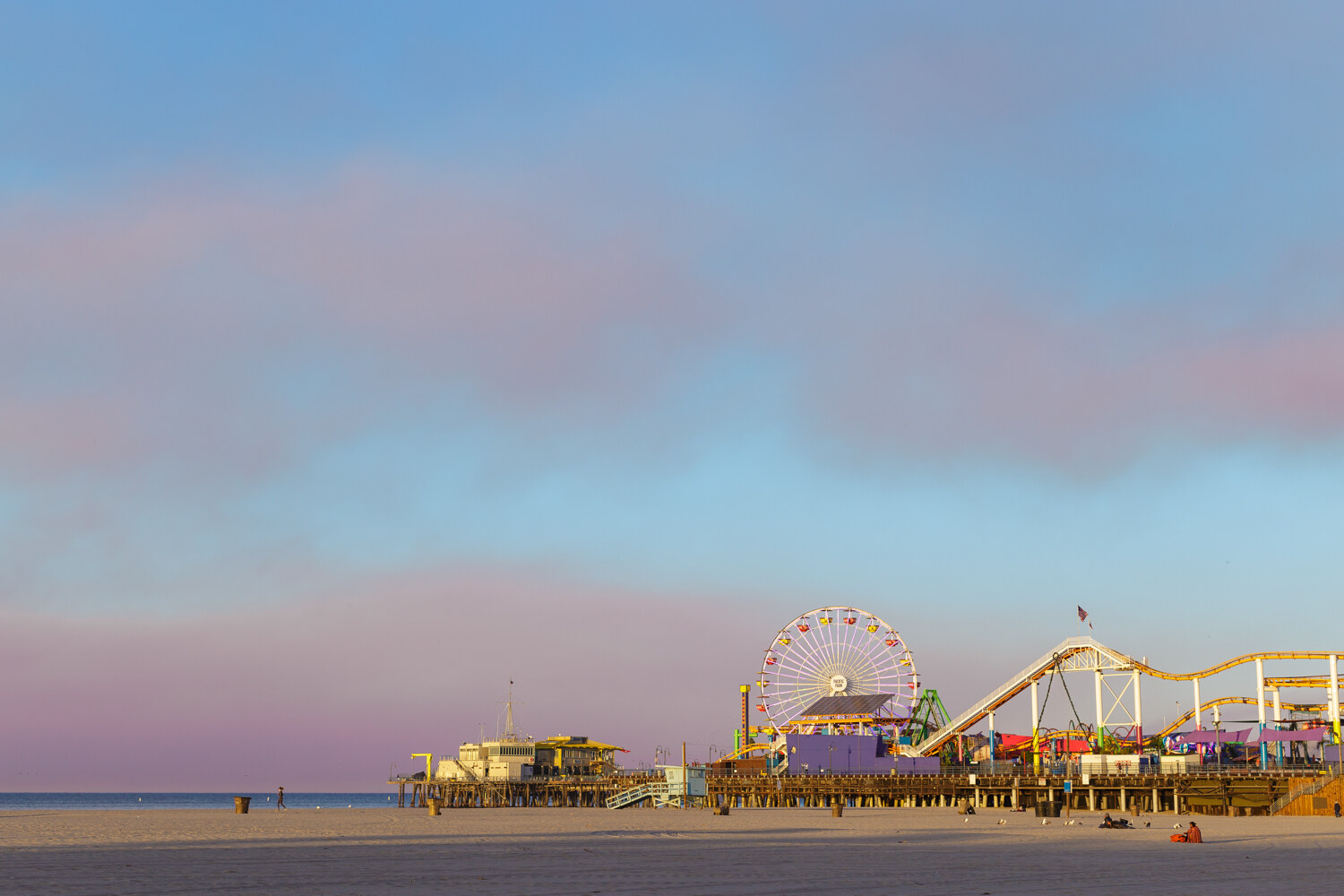 The Santa Monica Pier with its Ferris wheel and roller coaster seen across an empty stretch of beach under a pale blue and pink sky.