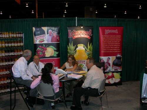 Four people sit around a small table with papers in a booth with green curtains, flanked by Lakewood Fresh Pressed banners and shelves of bottled juice.