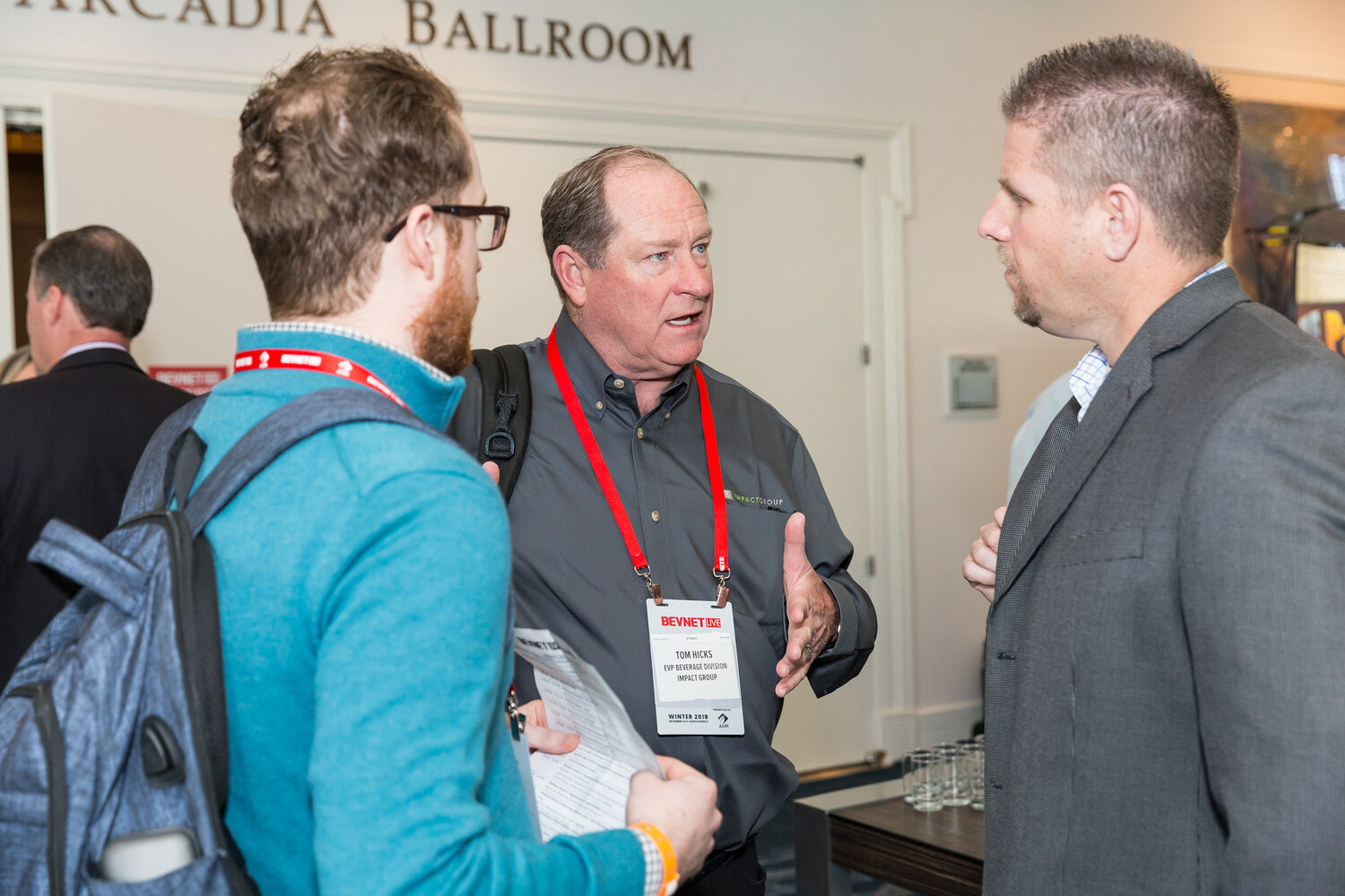 Three men with lanyards in conversation in front of a sign reading Arcadia Ballroom.
