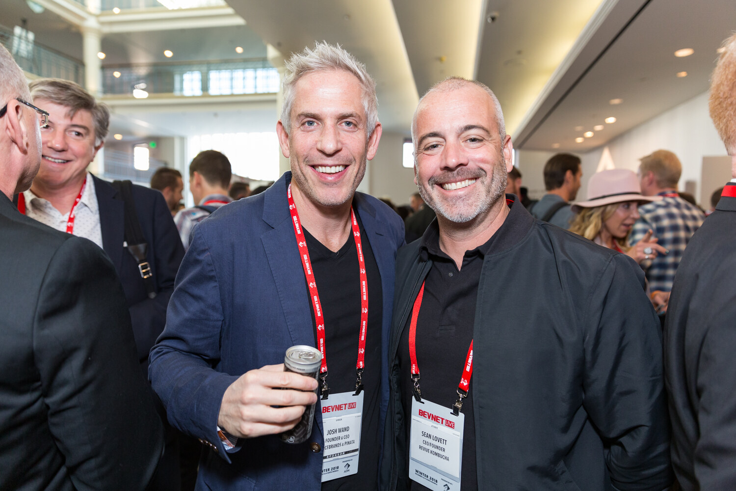 Two smiling men with red lanyards in a crowded ballroom.