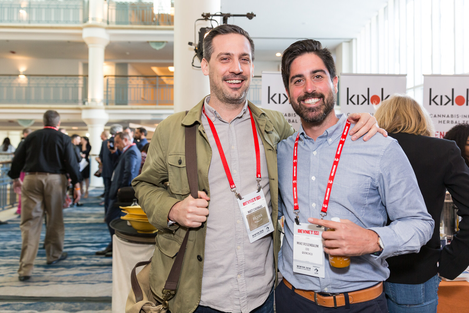 Two men with lanyards smiling together next to a Kikoko banner.