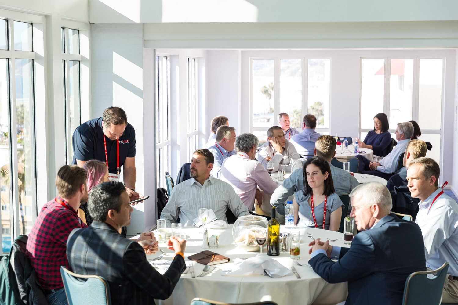 Round tables with attendees eating lunch in a room with large windows.