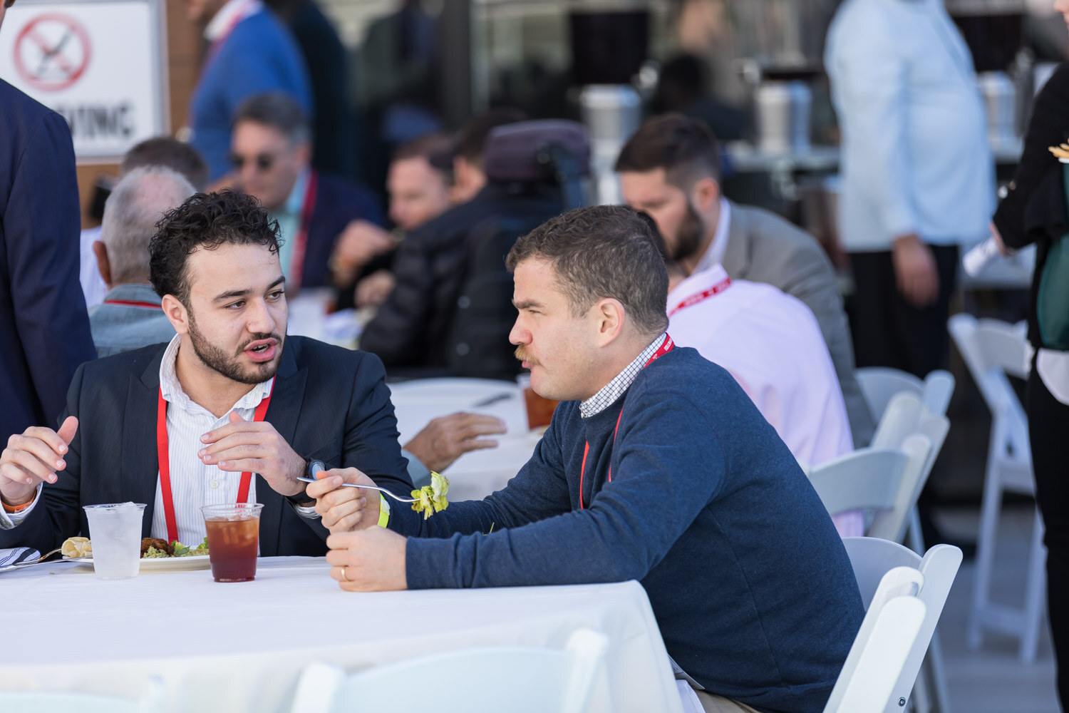 Two men in lanyards seated at a table talking over lunch.
