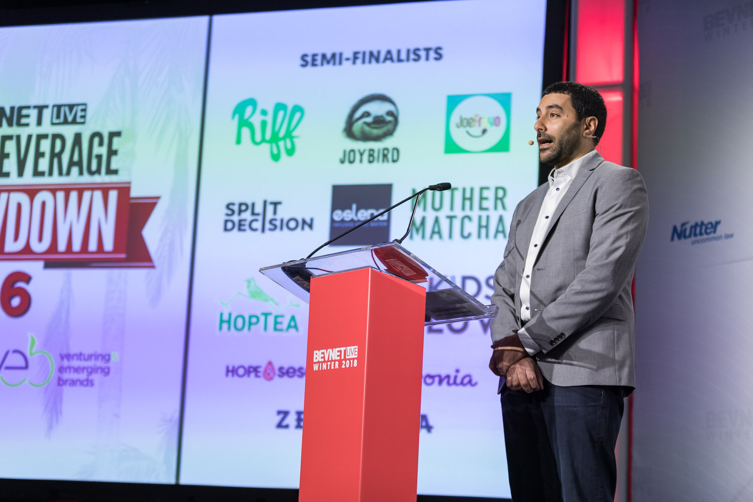 Man in a gray blazer at a podium beside a screen reading New Beverage Showdown 16 with semifinalist logos.