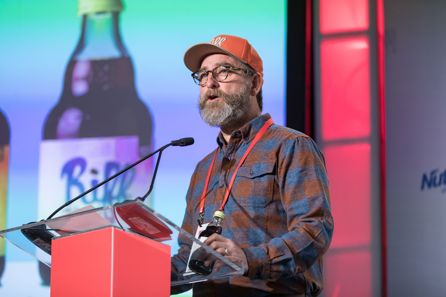 Man with a beard and orange cap at a podium in front of a large bottle on the screen.