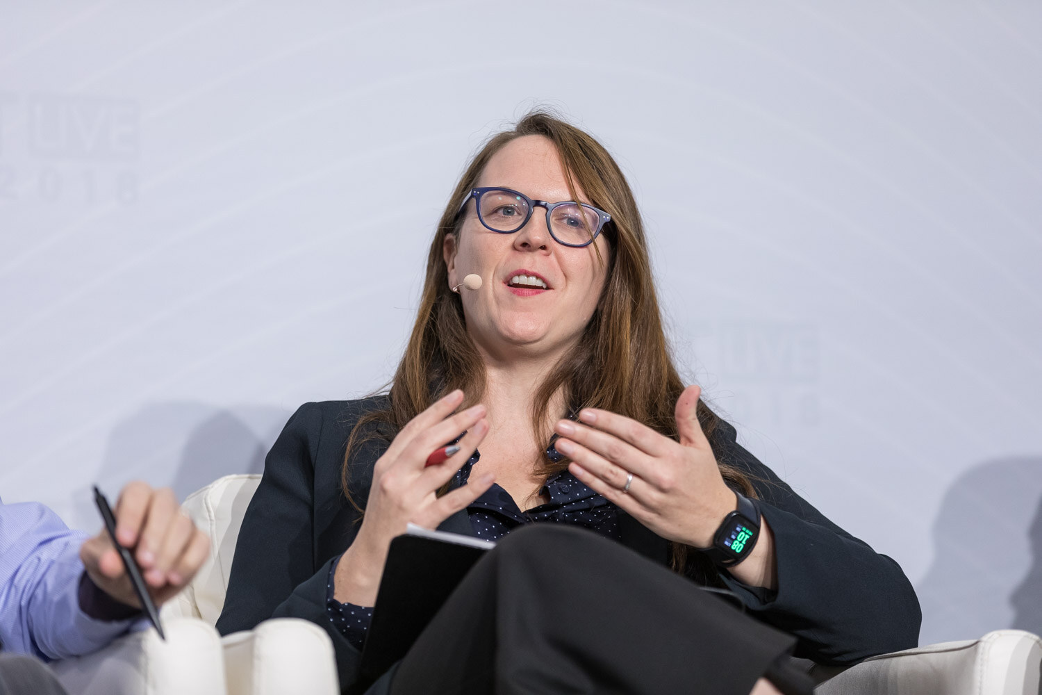 Woman with glasses and long hair gesturing while talking in a stage armchair.